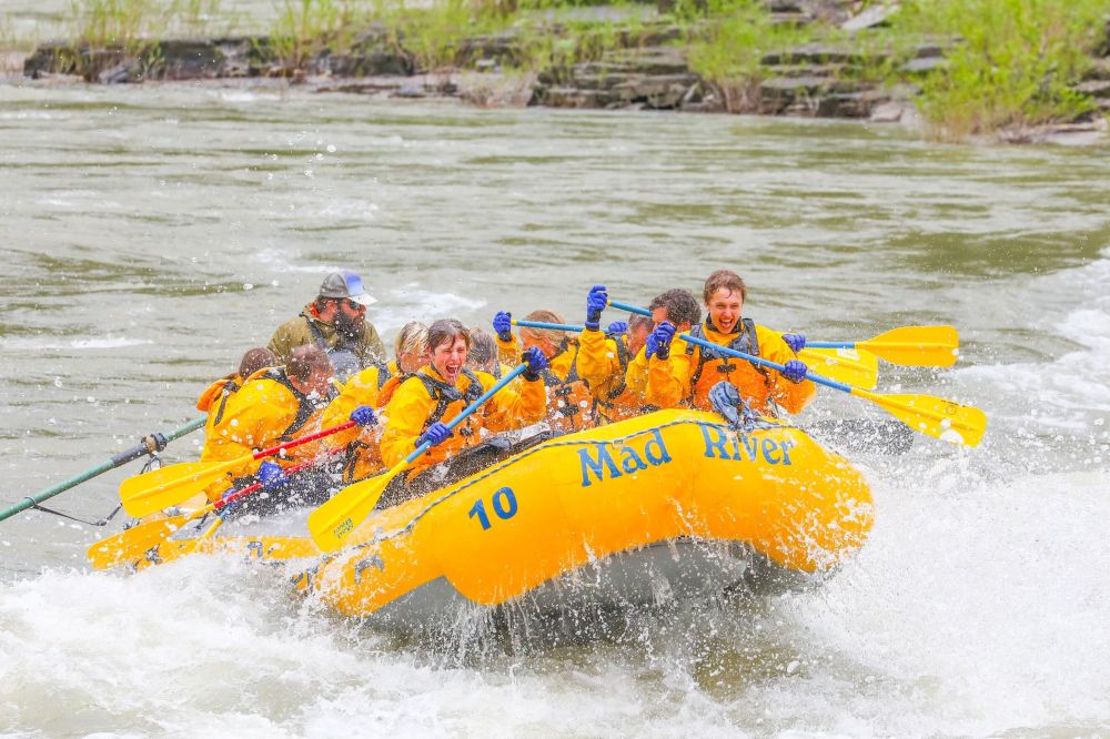 Group rafting on a river in a yellow boat labeled 'Mad River', splashing through rapids.